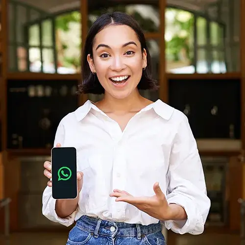 Mulher sorridente segurando um celular com o logo do WhatsApp na tela, vestindo camisa branca e jeans, em ambiente interno com fundo desfocado. Expressão de entusiasmo, como se estivesse indicando uma solução ou novidade no aplicativo.